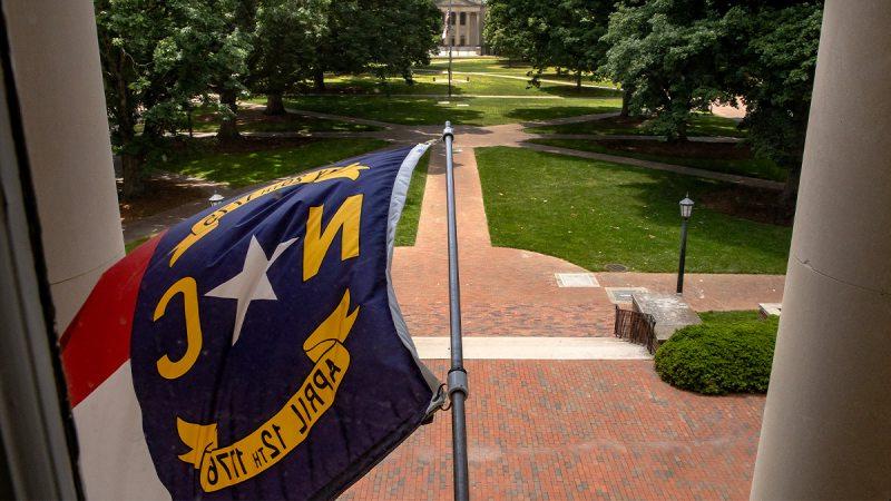 North Carolina state flag flying outside South Building on the campus of UNC-Chapel Hill with Polk Place seen in the background.